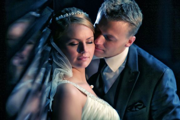 Ein Brautpaar in festlicher Kleidung vor einem schwach beleuchteten, romantischen Hintergrund, das Liebe und Eleganz verkörpert.