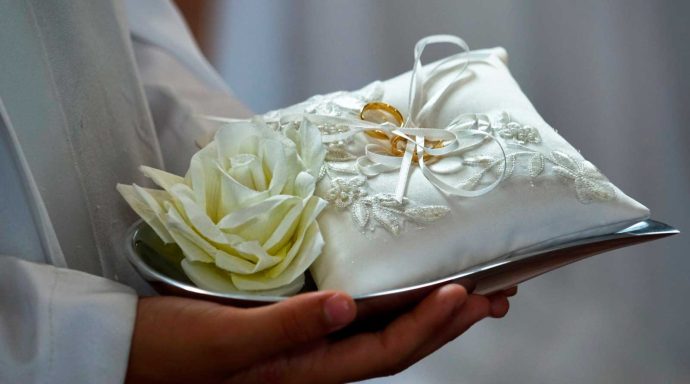 Zwei goldene Hochzeitsringe auf einer satinbezogenen Kissen, präsentiert von den Händen eines Pfarrers.
