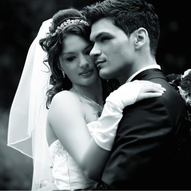 Das Bild zeigt ein romantisches Paar an ihrem Hochzeitstag vor einer Steinfassade, umgeben von Grün und sanften, warmen Lichtern.
