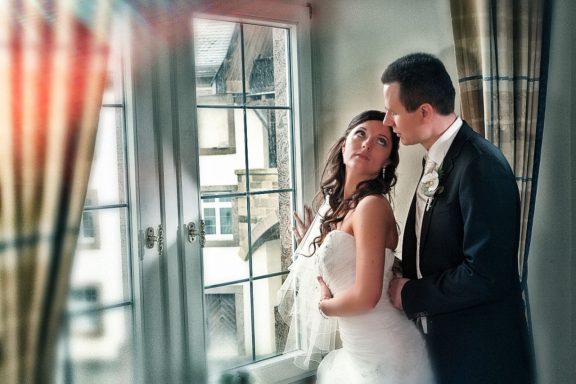 Ein Brautpaar umarmt sich liebevoll am Hochzeitstag, mit sanftem Licht und eleganter Kulisse durch ein großes Fenster.
