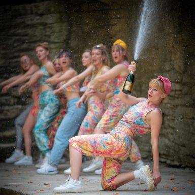 Eine Gruppe Frauen in bunten Outfits tanzt, eine spritzt Champagner aus der Flasche.