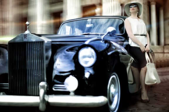 Eine Hochzeitsmoderatorin steht neben einem klassischen schwarzen Rolls-Royce auf einem Kopfsteinpflaster vor einem eleganten Gebäude mit Sä