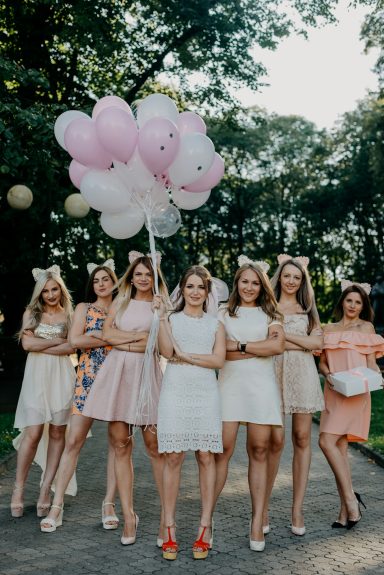 Gruppe junger Frauen in weißen Kleidern feiert im Freien, eine Frau in Pink, mit rosa und weißen Ballons.