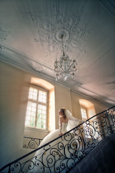 Eine Braut in einem weißen Hochzeitskleid schreitet elegant eine kunstvoll gestaltete Treppe hinauf, umgeben von warmem, natürlichem Licht.
