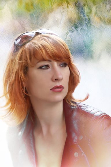 Person mit kurzem, orange-rotem Haar, in roter Lederjacke. Sonnenbrille auf dem Kopf vor unscharfem, grün-beigem Hintergrund.