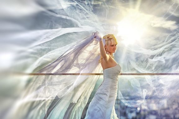 Frau in fließendem weißen Kleid mit transparenten Stoffen, umgeben von sanfter, leuchtender Atmosphäre vor dem Main Tower.  