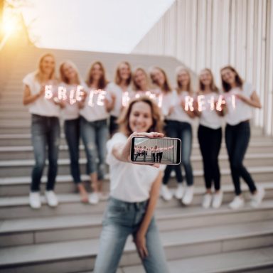 Eine junge Frau macht ein Selfie mit einem Smartphone, im Hintergrund eine Gruppe von sieben Frauen, die leuchtende Buchstaben halten, die "
