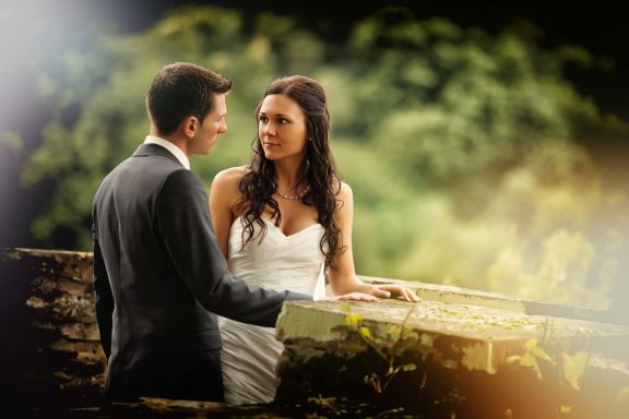 Das Bild zeigt ein romantisches Paar an ihrem Hochzeitstag vor einer Steinfassade, umgeben von Grün und sanften, warmen Lichtern.