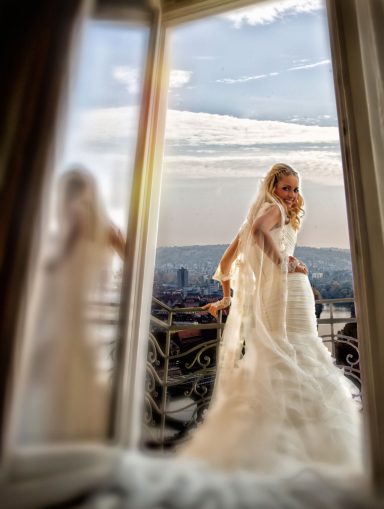 Braut in weißem Kleid steht an einem Fenster mit Blick auf den klaren Himmel und die Stadt.
