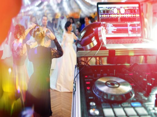 DJ-Setup mit Laptop und Mixer, ausgelassene Tanzenden im Hintergrund unter festlicher Beleuchtung bei einer Hochzeitsfeier.