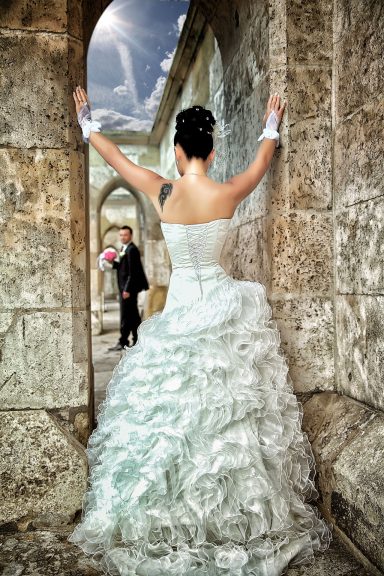  Eine Braut in einem weißen Hochzeitskleid mit vollem Zug steht vor einer steinernen Bogenarchitektur, mit festlicher Frisur, Rückenansicht.
