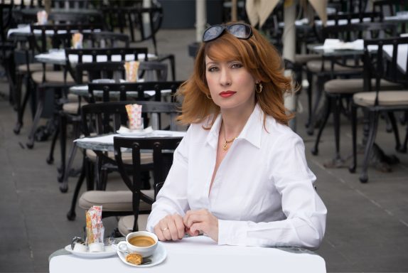 Hochzeit Moderatorin mit rotem Haar, in einer weißen Bluse, trinkt Kaffee im Freien an einem Café-Tisch.