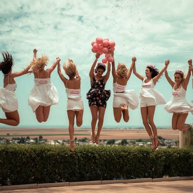 Sechs Frauen springen lachend in die Luft, die Braut in Weiß mit pinker Schärpe.