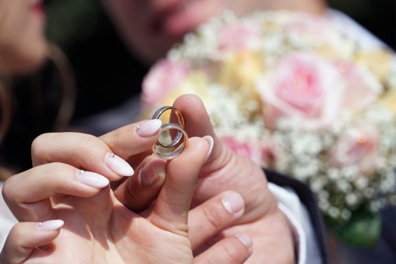  Zwei Hände halten goldene Hochzeitsringe, umgeben von einem verschwommenen Hintergrund mit rosa und weißen Blumen.