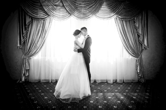 Brautpaar umarmt sich in formeller Hochzeitskleidung vor einem eleganten schwarzen Vorhang, die Atmosphäre ist romantisch und klassisch.